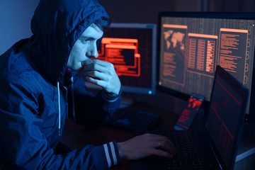 Wall Mural - Hacker in a hood thinking about the problem of hacking or malware infection on the background of screens with codes