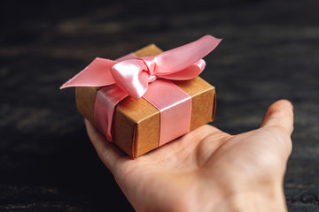 Wall Mural - Hand holding Holiday gift box Packed in crafting paper with pink ribbon on dark wooden background