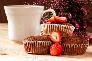 Chocolate muffins with strawberries, a cup of coffee and a pink heather in the background on a wooden table. Close-up.