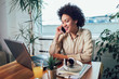 © Mediteraneo - Smiling young African female entrepreneur sitting at a desk in her home office working online with a laptop
