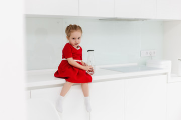 Cute clever little Caucasian girl in casual red dress holds a glass of water in her hands and drinks clear water, sits on a table in the white modern minimalistic kitchen. 