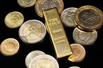 A macro image of an assortment of Turkish coins and a gold one ounce ingot on a reflective black background