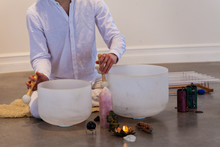 Man Playing Crystal Bowls. Man Dressed In White, Sitting On A Sheep Skin With A Close Up On His Hands Playing Crystal Bowls With Sacred Objects Displayed Like Stones, Candles And More