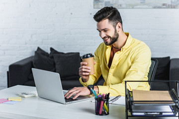 Wall Mural - Cheerful businessman holding paper cup and looking at laptop screen