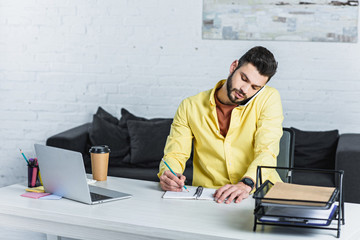 Wall Mural - Concentrated bearded businessman talking on smartphone and writing in notebook