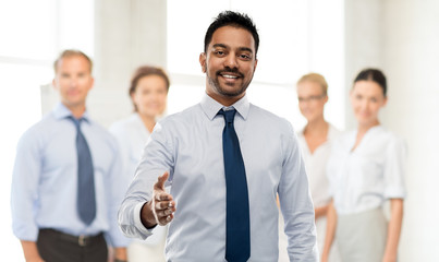 Wall Mural - business, office worker and people concept - smiling indian businessman stretching hand out for handshake over colleagues on background