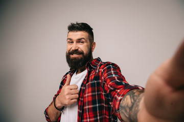 Wall Mural - smiling bearded man in checkered shirt showing thumb up while taking selfie, isolated on grey