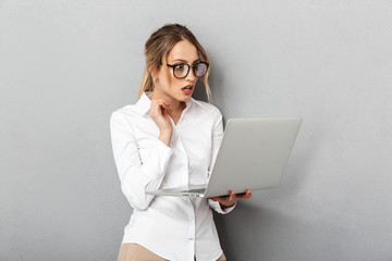 Sticker - Photo of attractive businesswoman wearing glasses standing and holding laptop in the office, isolated over gray background