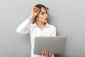 Sticker - Photo of european businesswoman wearing glasses standing and holding laptop in the office, isolated over gray background