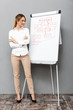 Full length image of joyful businesswoman in formal wear standing and making presentation using flipchart in the office, isolated over gray background