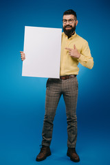 Wall Mural - bearded smiling man pointing at empty placard isolated on blue