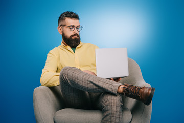 Wall Mural - bearded businessman sitting in armchair with laptop, isolated on blue