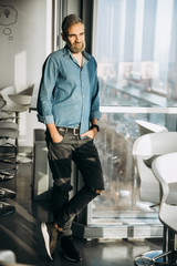 Wall Mural - Portrait of a modern handsome man with a beard in a blue shirt standing by the window indoors