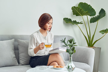 Sticker - Serious young Asian girl in blouse and skirt.sitting on sofa and drinking tea while reading book in living room