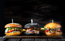 Three Different Burgers In A Line On A Wooden Board On A Dark Background