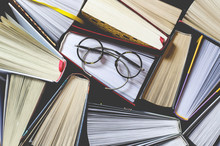 Many Multicolored Thick Open Books Stand On A Dark Background. On The Books Are Old Round Glasses And An Open Notebook With A Pencil.