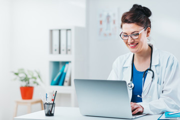 Wall Mural - Specialist looking at results from exam on laptop. Friendly woman doctor working on computer in her office