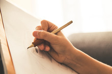 Close Up View Of A Young Hand Drawing On A White Sheet. Kid Hold A Black Wooden Pencil And Draw Something On A Warm Orange Light At Home. Children Writing On A Paper. Teen Drawing Freehand A Manga.