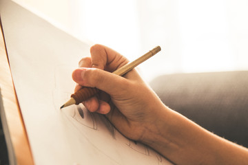 Close up view of a young hand drawing on a white sheet. Kid hold a black wooden pencil and draw something on a warm orange light at home. Children writing on a paper. Teen drawing freehand a manga.