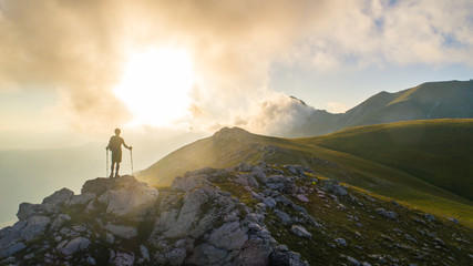 Wall Mural - young hiker man with backpack and walking poles, standing on peak of a mountain looking at sunset in