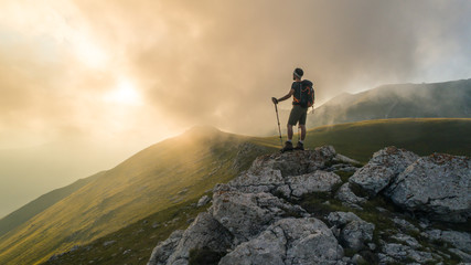 Wall Mural - young hiker man with backpack and walking poles, standing on peak of a mountain looking at sunset in