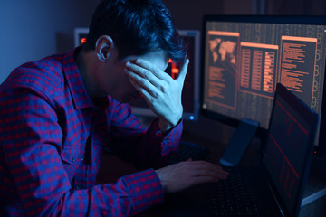 Wall Mural - Anonymous male hacker covers his face with hand while committing a crime to hack the system on the background of screens