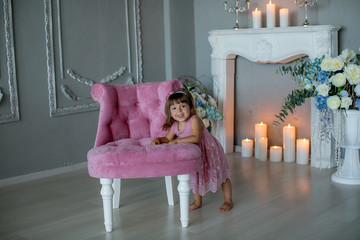 Smiling baby girl under 1 year old wearing stylish clothes sitting in vintage chair over white fireplace in room. Holiday season. Childhood.