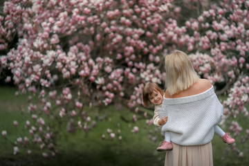 Portrait of happy joyful child in white clothes over tree flowers blossom background. Family playing together outside. Mom cheerfully hold little daughter Newborn spring concept