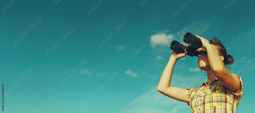 Beautiful Young Girl Looking Through Binoculars On Blue Sky Background. Travel Holidays Journey Concept - obrazy, fototapety, plakaty 