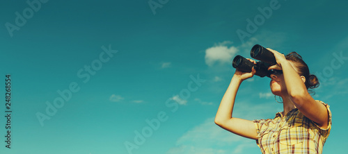 Cuadros en Lienzo Beautiful Young Girl Looking Through Binoculars On Blue Sky Background