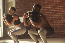 Young Mixed-race Fit Couple Doing Squats Exercise, With Heavy Sandbag In Gym. A Crossfit Sand Bag Is Advanced Level For Athletes Of Various Sports, Such As Crossfit Trainings And Contact Sports
