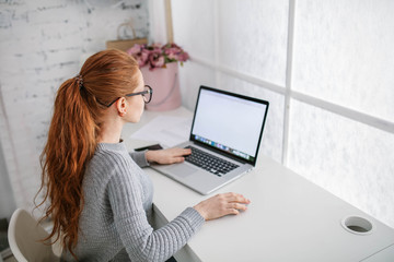 Wall Mural - Young beautiful woman with red hair, wearing glasses, working in the office, uses a laptop and mobile phone