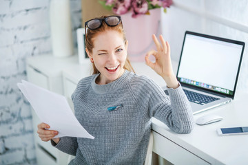 Wall Mural - Young beautiful woman with red hair, wearing glasses, working in the office, uses a laptop and mobile phone