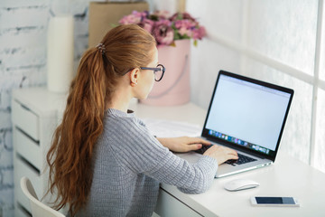 Wall Mural - Young beautiful woman with red hair, wearing glasses, working in the office, uses a laptop and mobile phone