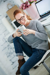 Wall Mural - Young beautiful woman with red hair, wearing glasses, working in the office, uses a laptop and mobile phone