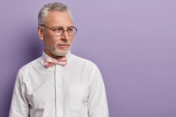 Wall Mural - Sideways shot of thoughtful grey haired man with thick beard, wears spectacles, elegant shirt, focused aside, deep in thoughts, thinks up next step, isolated over purple studio wall with empty space