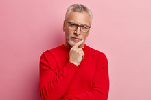 Headshot Of Handsome Grey Haired Man Holds Chin, Dressed In Red Sweater, Looks Directly At Camera, Has Thoughtful Expression, Poses Over Rosy Background. Pensive Grandfather Has Attentive Gaze