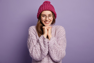 Wall Mural - Studio shot of pleased smiling lady keeps hands together under chin, wears warm hat and loose sweater, isolated over purple background. People, good emmotions, feelings concept. Woman ready for winter