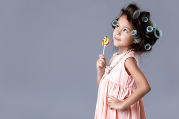 Wall Mural - Funny little child girl in pink dress and hair curlers holding lollipop on gray background.