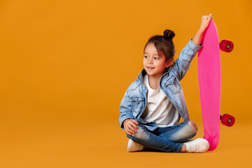 stylish little child girl with skateboard in jeans clothes on orange background
