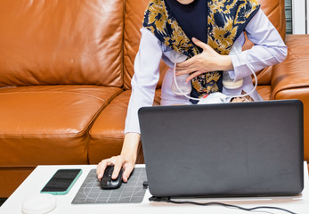 Busy young modern mother working with laptop while using automatically electric breast pump feeding for her baby.