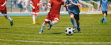 Football Soccer Players Running With Ball. Footballers Kicking Football Match. Young Soccer Players Running After The Ball. Kids In Soccer Red And Blue Uniforms. Soccer Stadium In The Background