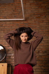 young, thin and attractive girl student dressed in casual clothes, red pants and a brown jacket, poses for a photo opposite the stone wall in the photo studio