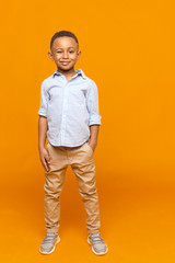 Wall Mural - Fashion, style, children's wear and childhood concept. Full length picture of cool handsome dark skinned model of school age winking at camera, posing at orange wall with copyspace for your text