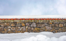 Stone Wall Covered By Fresh Snow In Winter. A Clear Sky With Soft White Clouds Appears In The Background