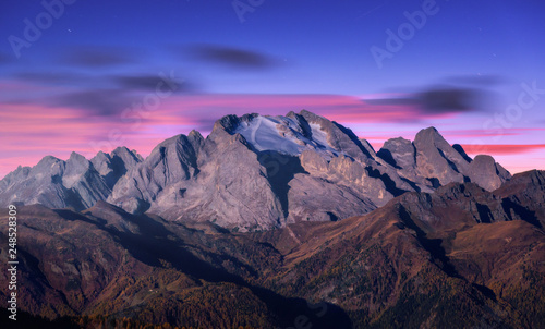 Mountain peak lighted by moonlight in autumn at night in Dolomites, Italy. Beautiful landscape with mountains, forest on hills, purple sky with pink clouds, stars at dusk. Italian alps. High rocks
