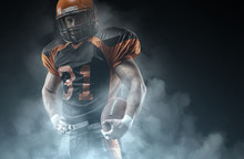 American Football Player On A Dark Background In Smoke In Black And Orange Equipment.