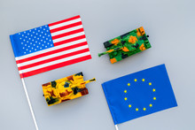 War, Confrontation Concept. European Union, USA. Tanks Toy Near European And American Flag On Grey Background Top View