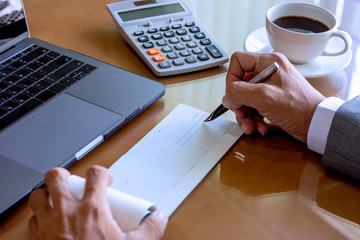 Wall Mural - Business man hand writing and signing cheque book with laptop computer calculator and cup of coffee on the wooden table at modern home office.	