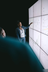 Black african businesswoman giving a presentation at a business conference. Pointing at a large video screen with charts and graphs next to her in a dark room